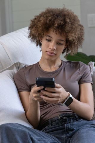 African american woman relaxing on sofa using smartphone and smartwatch for messaging
