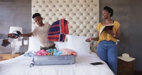 African American Family Packing for Vacation on Bed, Mother Checking Notebook, Son Tossing Towel