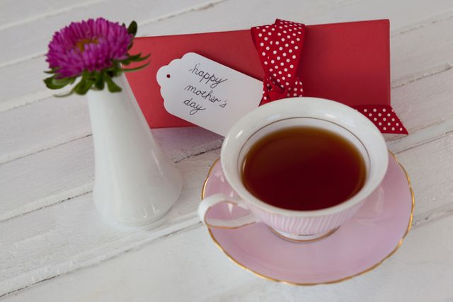 Perfect for Mother's Day promotions, greeting cards, and social media posts celebrating mothers. The image features a cup of black tea in a pink cup and saucer, a red gift with a polka dot ribbon, and a small flower in a white vase, creating a warm and festive atmosphere.