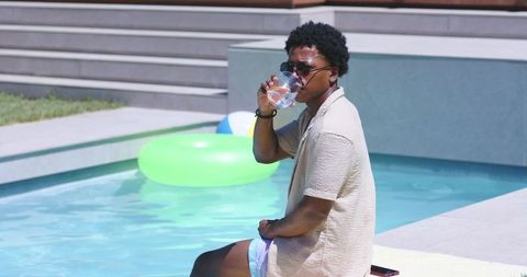 Man relaxing by modern backyard pool sipping drink in sunglasses and summer shirt