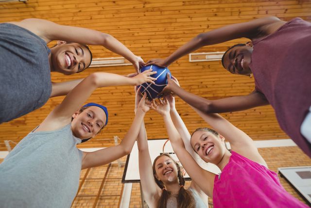 Group of diverse high school kids smiling and holding a basketball together in a gym. Perfect for promoting teamwork, youth sports programs, school activities, and fitness initiatives. Ideal for educational materials, sports event promotions, and community engagement campaigns.