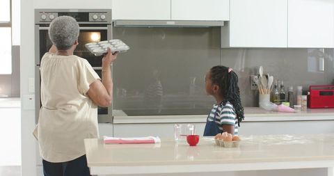 Senior african grandmother placing muffin tray in oven while granddaughter watches