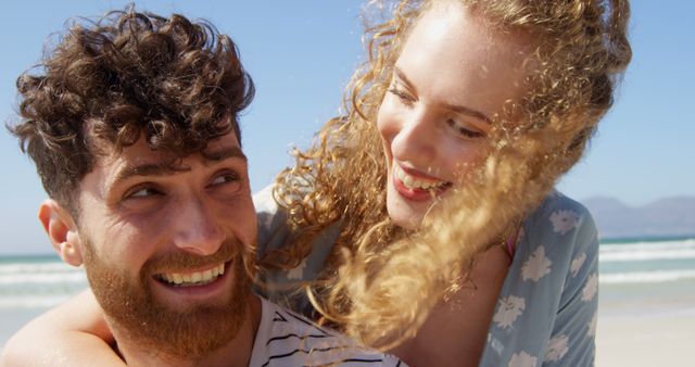 This image captures a young Caucasian couple enjoying a loving moment on a sunny beach. Their joyful expressions and close interaction highlight themes of happiness, bonding, and fun in an outdoor setting. Ideal for use in advertising for travel agencies, vacation packages, couples' retreats, or romantic getaways. Great for illustrating articles on relationships, summer, or beach vacations.