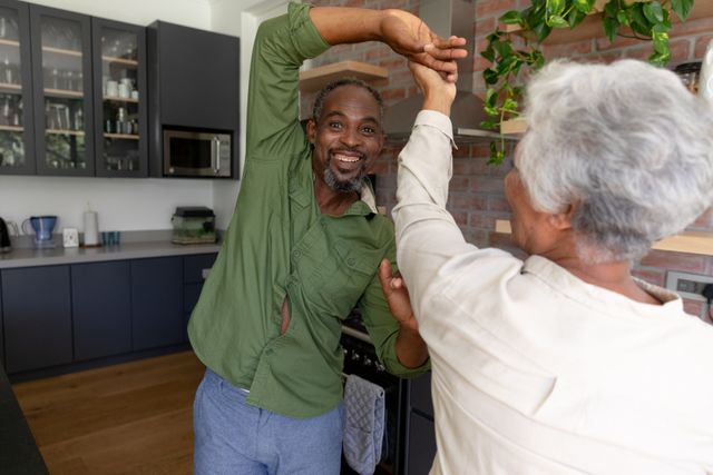 Elderly African American couple enjoying a joyful moment dancing in their home kitchen. Ideal for concepts related to love, togetherness, happiness, and life during quarantine. Can be used in articles about senior lifestyle, relationships, and coping with isolation during the Covid-19 pandemic.
