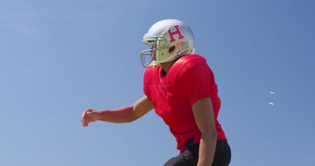 Football player dressed in red jersey and silver helmet is captured in an outdoor setting with a clear, blue sky in the background. Ideal for promoting sports apparel, fitness, American football events, team activities, motivational themes, and high-energy sports scenes.