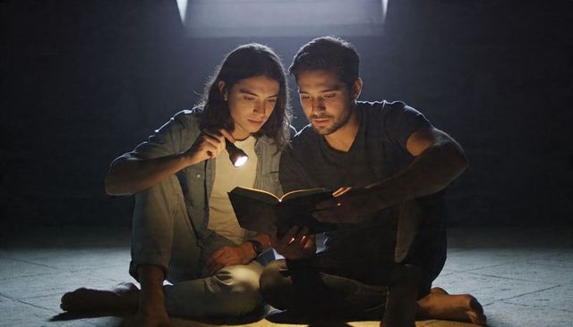 Two men reading by flashlight under skylight in dim basement, intimate nighttime scene