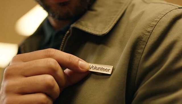 Bearded man adjusting volunteer badge on olive jacket during community event