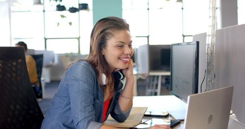 Young woman smiling while working at laptop in modern open-plan office with headset
