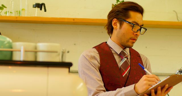 Young male doctor with glasses writing notes on a clipboard in a medical office. Wearing a formal outfit with a striped tie and red vest. This image could be used for themes around healthcare, professional environments, medical staff, or diligent work.