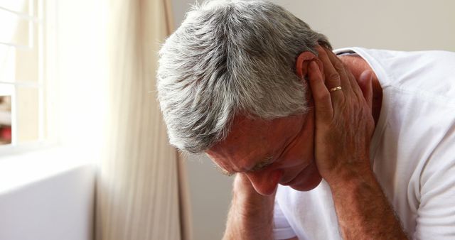 A senior man, with white hair, holding his head while sitting at home, seemingly experiencing a strong headache or stress-related discomfort. Great for illustrating issues related to elderly health, pain management, stress, or general well-being. Ideal for use in health articles, medical resources, caregiver support materials, and wellness guides.
