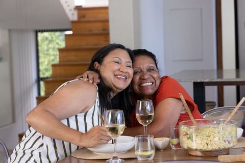 Multigenerational women hugging at sunlit dining table with pasta salad and wine, candid