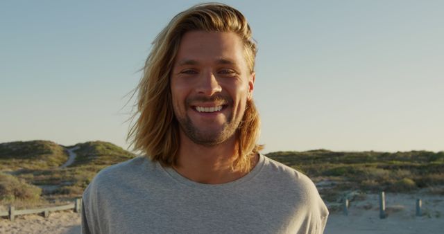 Portrait of a smiling young man with long hair enjoying a day at the beach. The background shows a serene beach setting with sand dunes and a clear sky. Perfect for lifestyle, travel, summer activities, and casual fashion themes.