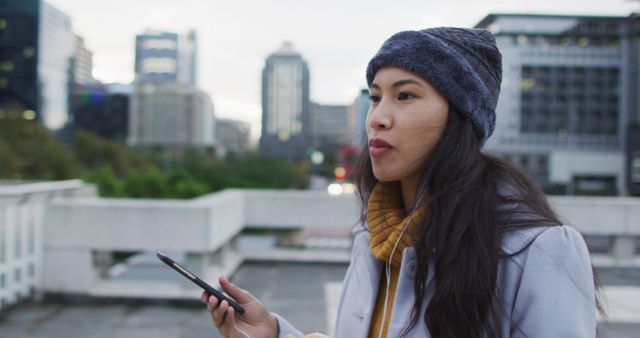Asian woman in urban cityscape using smartphone, dressed in autumn attire, connecting with technology. Useful for depicting urban lifestyle, technology usage, modern communication, or seasonal outdoor scenarios.