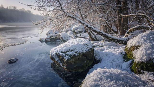 Snow-topped boulders reflecting on icy river with mossy rocks and misty winter shoreline