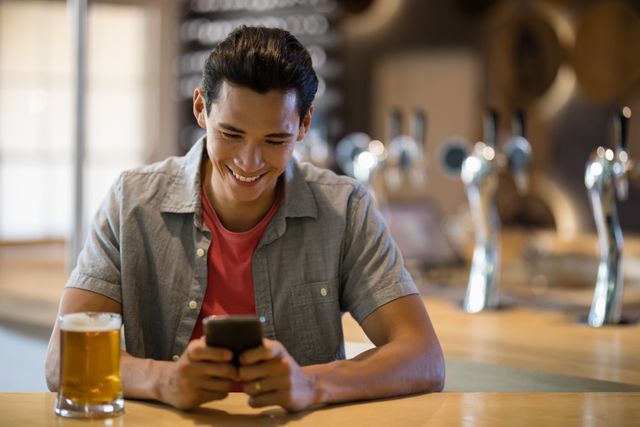 Young man sitting at a bar counter, smiling while looking at his mobile phone. A glass of beer is placed on the counter. Ideal for use in advertisements related to social media, technology, leisure activities, or bar and restaurant promotions.