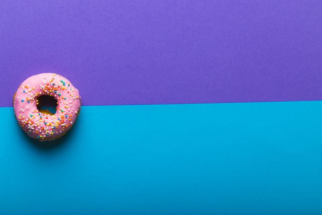 Pink donut with colorful sprinkles placed on a vibrant two-tone purple and blue background. Ideal for use in advertisements, social media posts, or blog articles related to desserts, unhealthy eating, or colorful food presentations. The minimalistic design and ample copy space make it perfect for promotional materials and creative projects.