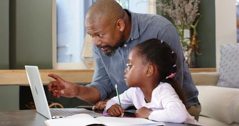 Father tutoring daughter at home using laptop and workbook for remote learning