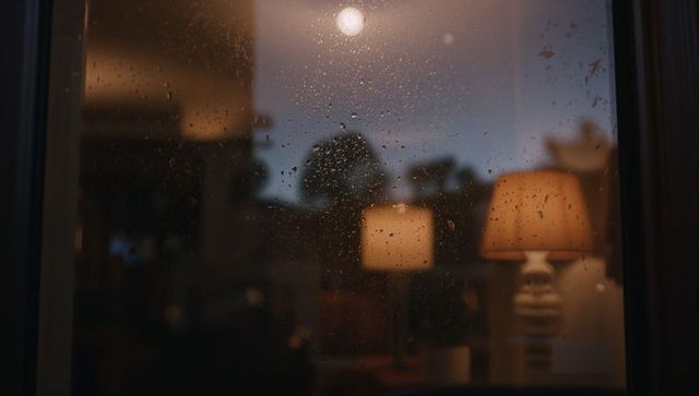 Rain-Soaked Windowpane Glimmering at Dusk Showing Warm Cozy Living Room Lamp Glow