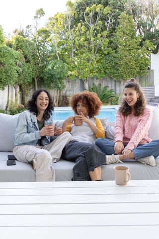 Diverse female friends relaxing on outdoor sofa by pool holding coffee mugs and laughing