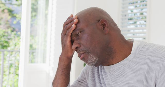 Elderly African American man holding head in distress indoors. He appears worried and stressed, conveying a range of emotions. Suitable for articles and campaigns on mental health, stress, emotional well-being, senior health care, and personal challenges.