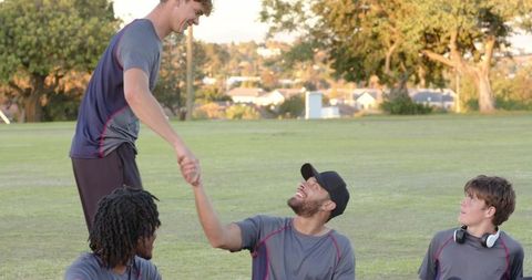 Young men helping teammate up on grassy field at sunset showing teamwork and friendship