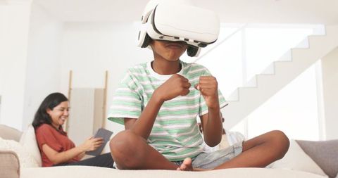 Child wearing VR headset playing virtual boxing on sofa while parent relaxes with tablet