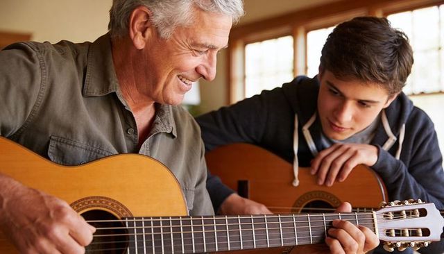 Senior teaching teen guitar at home — intergenerational music lesson in warm daylight