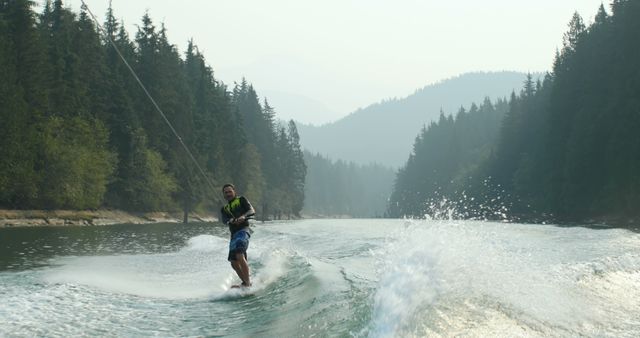 Man wakeboarding on serene lake surrounded by dense pine forest on a foggy day. Suitable for depicting outdoor adventures, water sports, nature escapes, and active lifestyles. Ideal for travel brochures, adventure magazines, sports advertisements, and tourism websites.