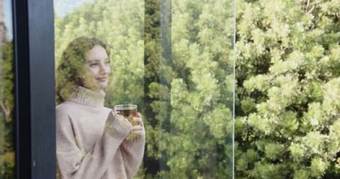 Contemplative Woman Holding Glass Mug by Tall Window with Nature View