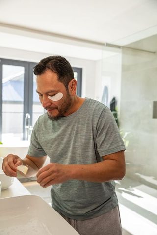 Mature asian man applying under-eye patches during bright morning skincare routine at sink