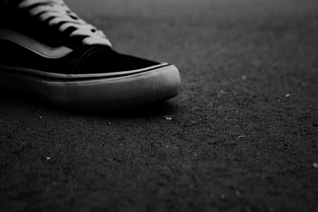 This black and white image shows a close-up of a black sneaker on an asphalt ground. The minimalistic composition and monochrome filter add a dramatic effect, ideal for use in urban lifestyle blogs, fashion websites, or promotions emphasizing simplicity and style.