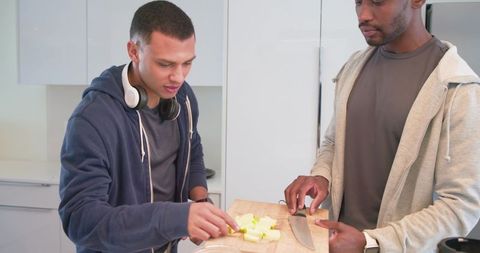 Diverse male friends chopping fresh vegetables on wooden cutting board in bright modern kitchen