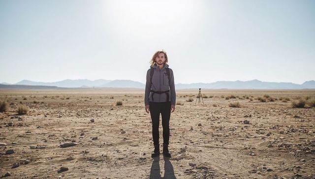 Solo hiker standing on barren desert plain with backpack casting long shadow and tripod