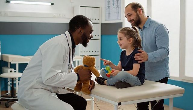 Smiling pediatrician playing with child and father on clinic exam table with teddy bear