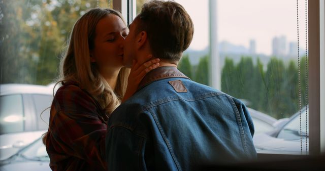 Young couple sharing a kiss near window with greenery and parked cars outside. Presentation of love and intimacy in a casual setting. Ideal for relationship articles, romantic product promotions, greeting cards, social media posts about love and romance.
