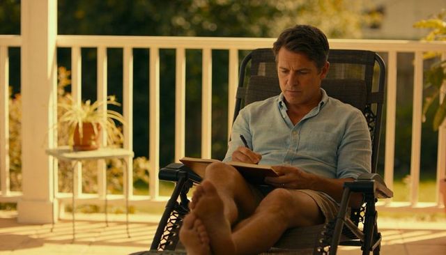 Middle-aged man writing in journal on sunny porch recliner barefoot, relaxed summer lifestyle