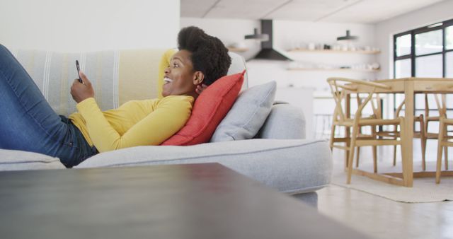 Young woman comfortably lounging on couch, using smartphone. Modern and bright living room. Perfect for themes of relaxation, home life, casual lifestyle, and use of technology in daily routines.