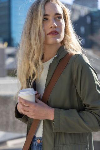 Young woman holding takeaway coffee in city plaza wearing olive jacket