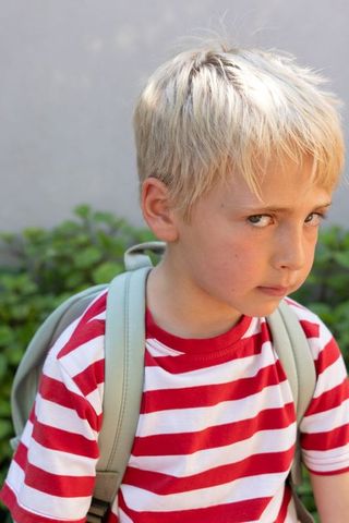 Blond boy standing in backyard wearing backpack and red striped shirt giving guarded look