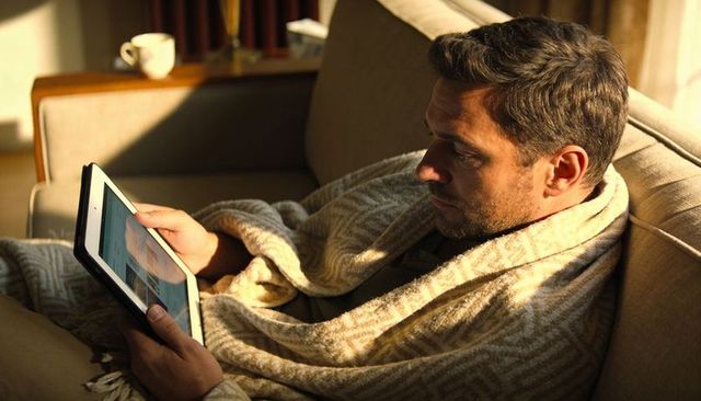Man relaxing on sofa using tablet while snuggling in cozy patterned blanket in warm sunlight