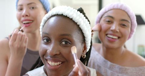 Multiethnic friends applying makeup together at home vanity, smiling and sharing tips