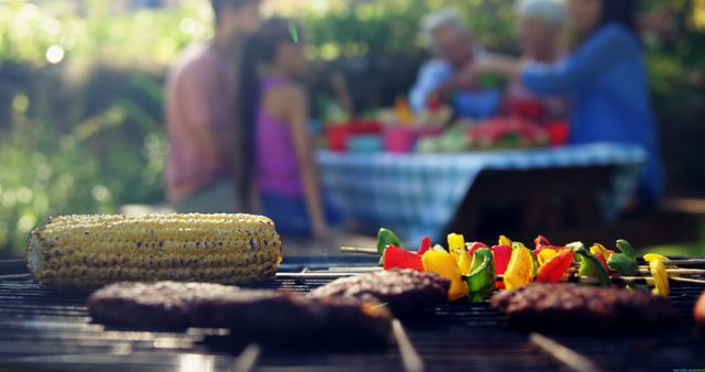 Foreground shows a barbecue grill with corn, burgers, and skewered vegetables, with copy space. In the background, a diverse family enjoys a meal together outdoors, capturing a moment of leisure and togetherness.