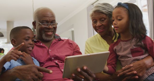 Senior couple spending time with grandchildren using a digital tablet at home. The grandparents are teaching and bonding with the children, who are engaged and smiling. This image captures warm family moments and can be used for concepts related to family bonding, technology use, intergenerational relationships, and happy home life.