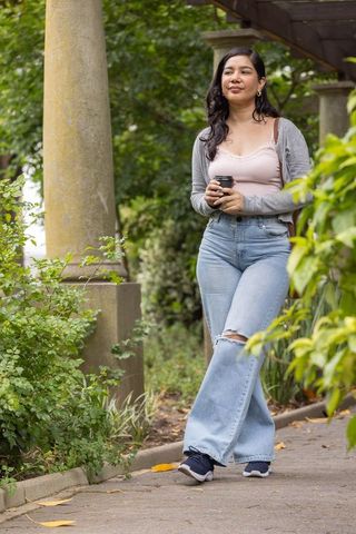 Woman strolling in park holding takeaway coffee cup by stone column pergola in denim