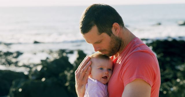 Caucasian father holding happy baby to his chest on sunny beach by the sea, copy space. Fatherhood, care, wellbeing, vacations, nature and travel, unaltered.