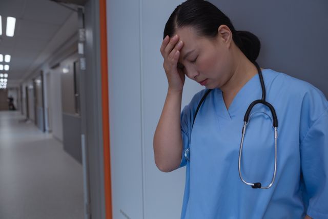 This image depicts a female doctor in blue scrubs, leaning against a wall in a hospital corridor with her hand on her forehead, indicating stress or fatigue. It can be used in articles or presentations about healthcare worker burnout, mental health in the medical field, or the challenges faced by medical professionals. It is also suitable for illustrating the human side of healthcare and the emotional toll of working in a hospital.