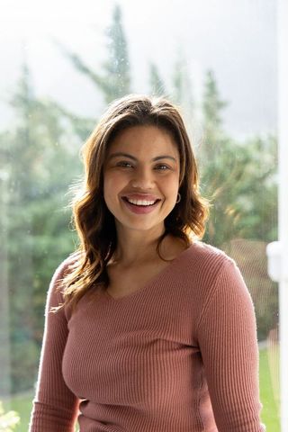 Smiling woman standing by sunlit floor-to-ceiling window wearing ribbed sweater