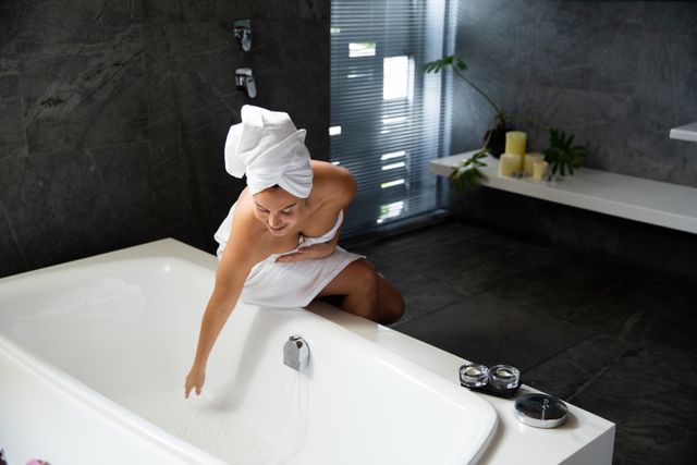 Front view of a Caucasian woman wearing a bath towel and with her hair wrapped in a towel, sitting on the edge of the bath and touching the water in a modern bathroom