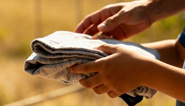 Parent handing folded light-gray t-shirts to child in sunlit backyard close-up