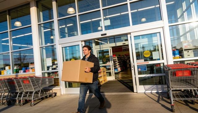 Man carrying large cardboard box exiting supermarket entrance with shopping carts nearby